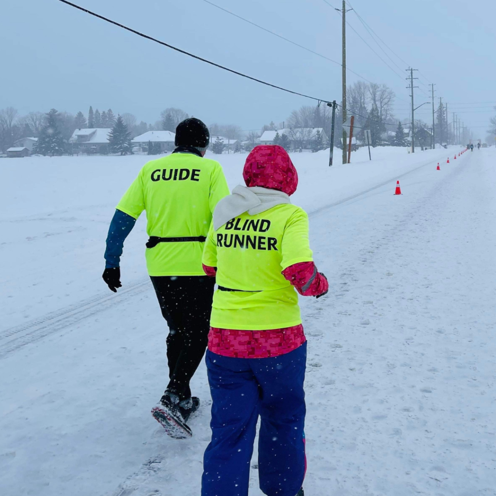 Two runners competing in a 10K race in the snow.  Both in brightly coloured shirts, one with Guide written on the back, the other with Blind Runner.