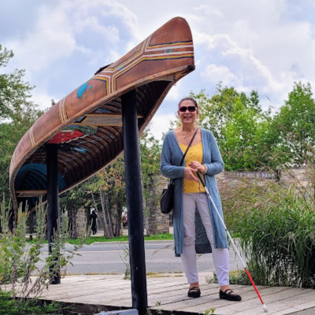 Smiling Woman walking with a white cane in a park.  Statue of a Canoe to her right.