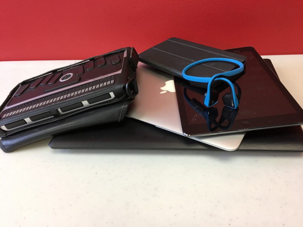 A pile of various technology including a braille display, bone conductive headphones, a laptop, an iPad, and tablet.