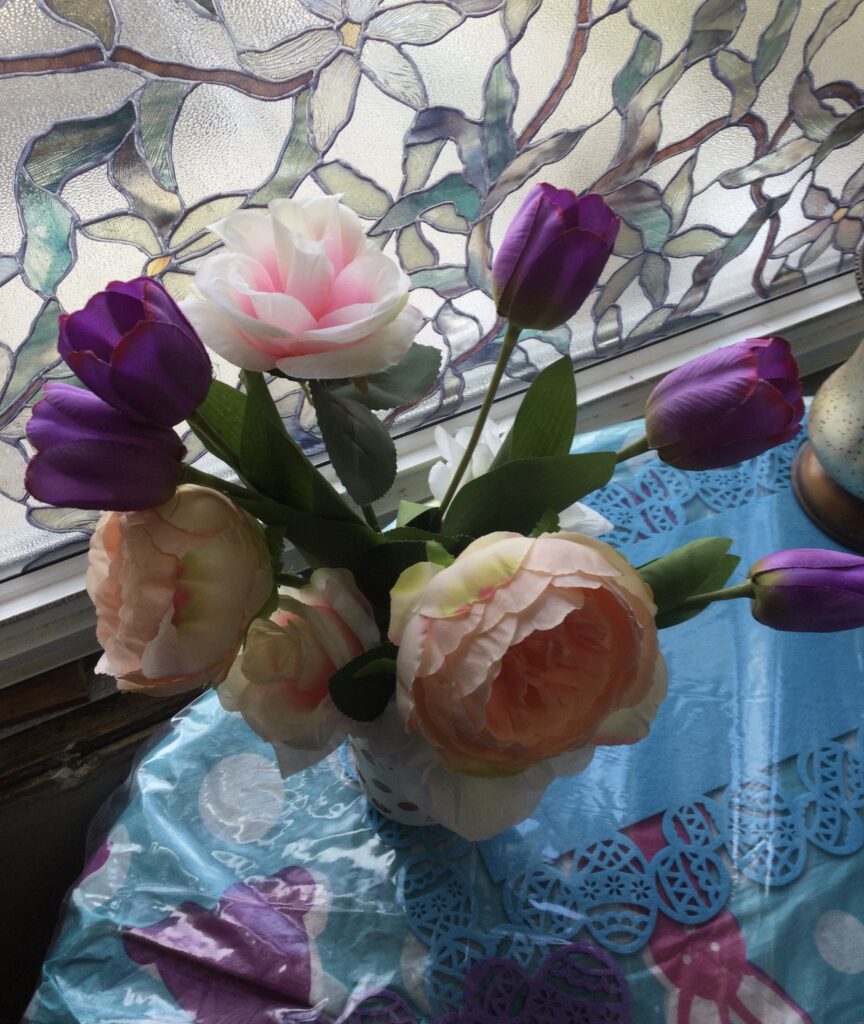 Flower arrangement, of pink and purple flowers on a small table on front of a window that appears partially.