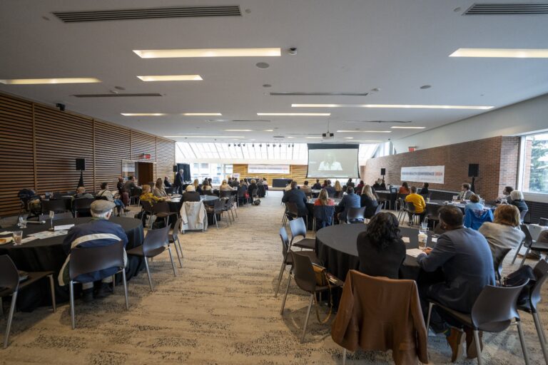 A large conference room with round tables set up for attendees, who are seated and watching a presentation at the front. A projector screen displays a speaker. The room is well-lit with natural light from large windows on the far wall. Attendees are engaged in the session, with several people seated in groups at each table.