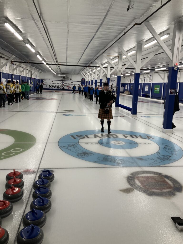 A curling rink with players lined up along the ice, dressed in team uniforms, preparing for an event. A bagpiper in a traditional Scottish kilt and black jacket stands on the ice, playing the bagpipes. The curling stones are arranged in two rows, with red and blue handles. The ice surface features logos and sponsor markings, including a prominent 'Island Folk Cider House' logo on one of the houses. The venue has a high ceiling with bright overhead lighting, white beams, and banners along the walls. The scoreboard at the far end displays 'Sydney,' indicating the location.