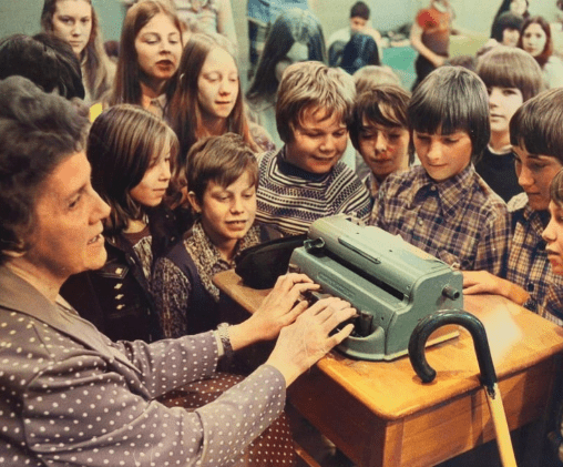 Historic photo of a woman demonstrating a braille typewriter to a group of children gathered around her.