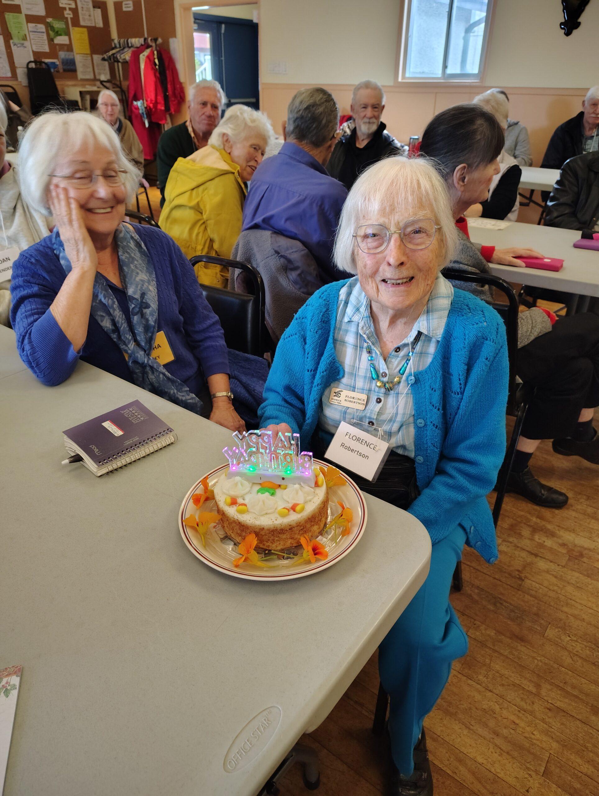 A photo of Florence Robertson smiling at the camera. She is sitting at a table next to her friend with a birthday cake in front of her.
