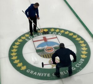 A photo of two people across from each other curling. They are standig above the Edmonton Granite Curling Club logo which is shown on the ice.