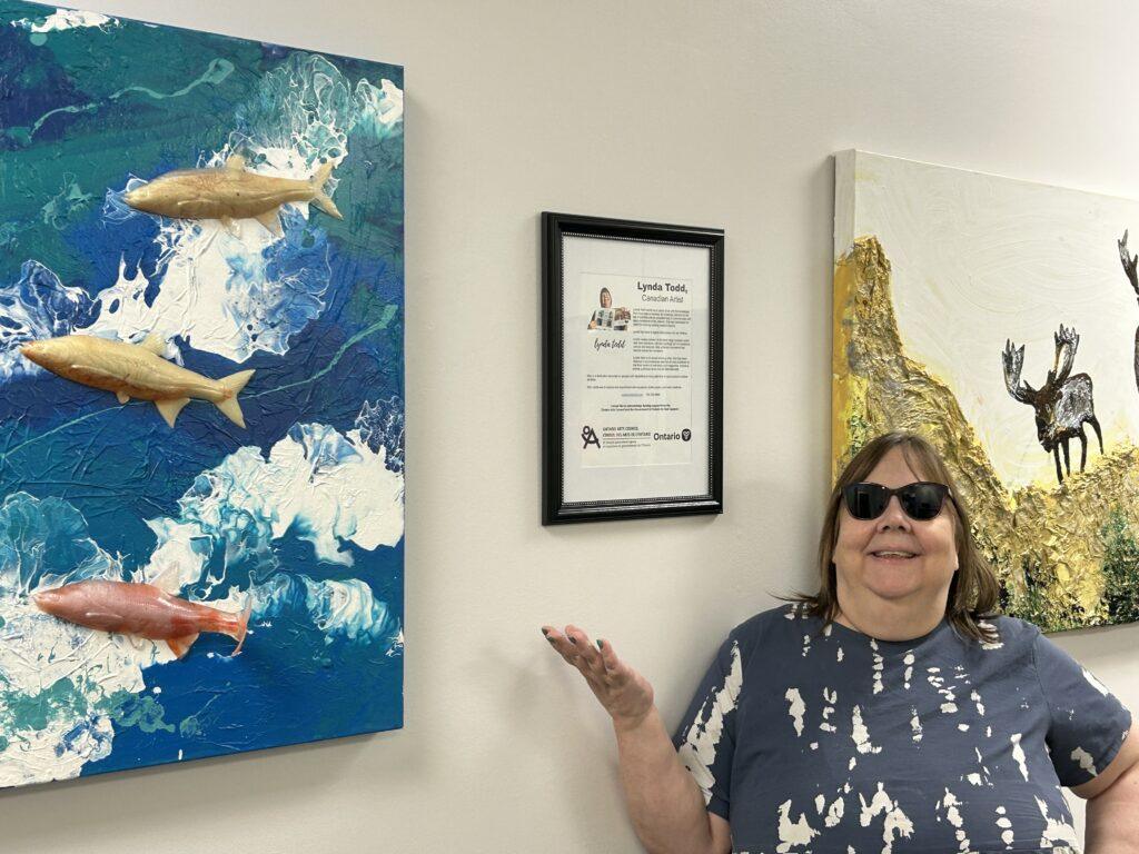 The picture shows Lynda Todd, wearing black sunglasses and a navy and white shirt, standing beside her artwork on a wall and gesturing toward it. The canvas on the left depicts three fish swimming in ocean water, and the canvas on the right shows a moose above a white background with golden scenery below.