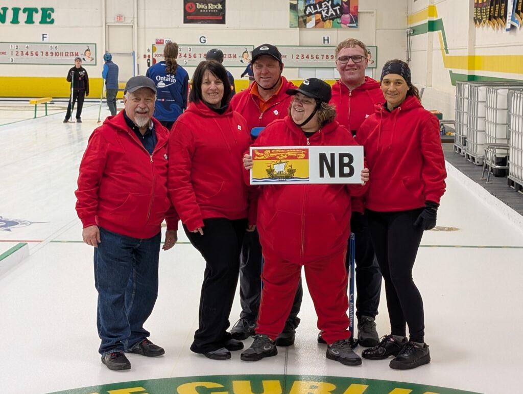 The picture shows players from Team New Brunswick standing side by side on the ice in a curling rink, posing for the camera. They are wearing their red team jackets. One player, Natalie Fougère, is holding a sign that shows the New Brunswick flag on the left and “NB” on the right.