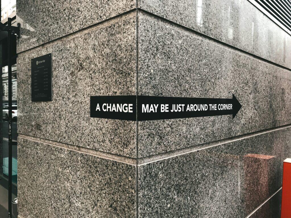 The picture shows the corner of a grey wall in a building with a black strip running across the center. The strip reads “A change may be just around the corner” in capital white letters.