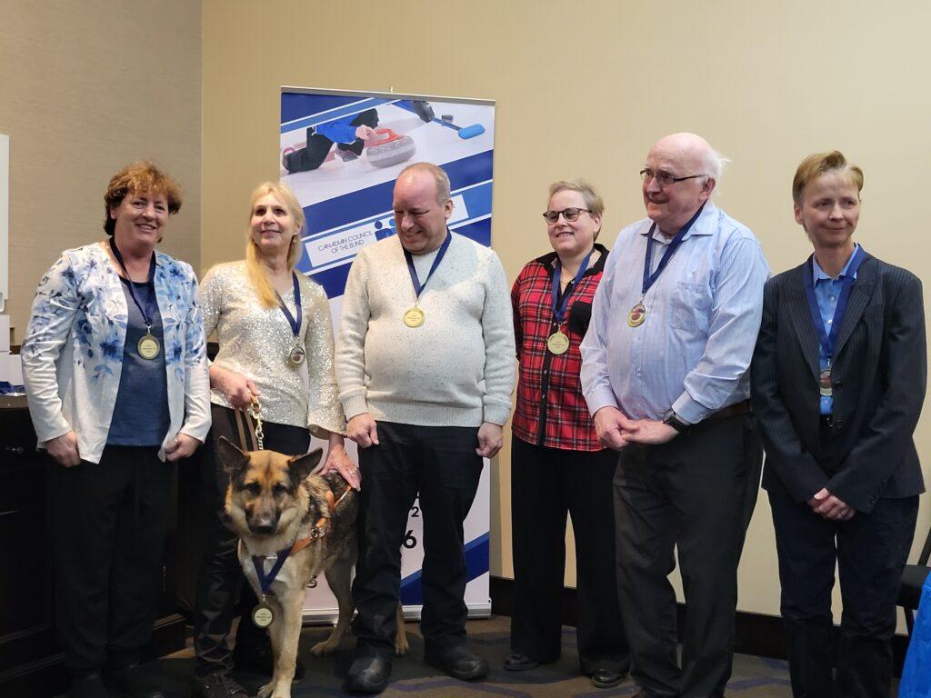 Players from Team Alberta, champions of the 2026 CVICC, stand in a room posing for the camera. Natalie Morin, second on the left, is standing with her guide dog in front of her. Each player wears their gold medal ribbons.