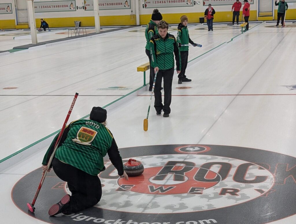 Team Saskatchewan players in green jackets are curling on the ice. One player is delivering a curling stone from a kneeling position, while teammates ahead can be seen holding brooms, with one ready to sweep.
