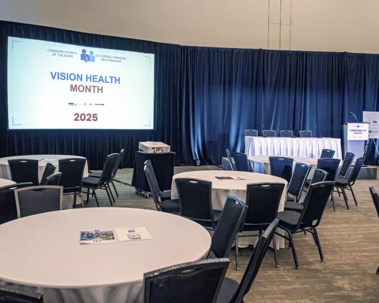 An image of the CCB Vision Month Conference 2025 event space. The image was taken before the conference had began and therefore there are no people present. The space features white linen round tables and a presentation screen with the conference logo prominently displayed.