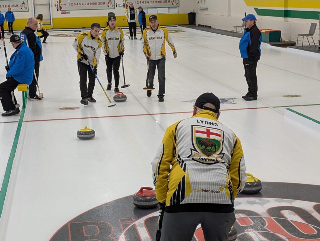 Team Manitoba players in yellow and white jackets sweep the ice while Team Alberta players in black and blue jackets watch.