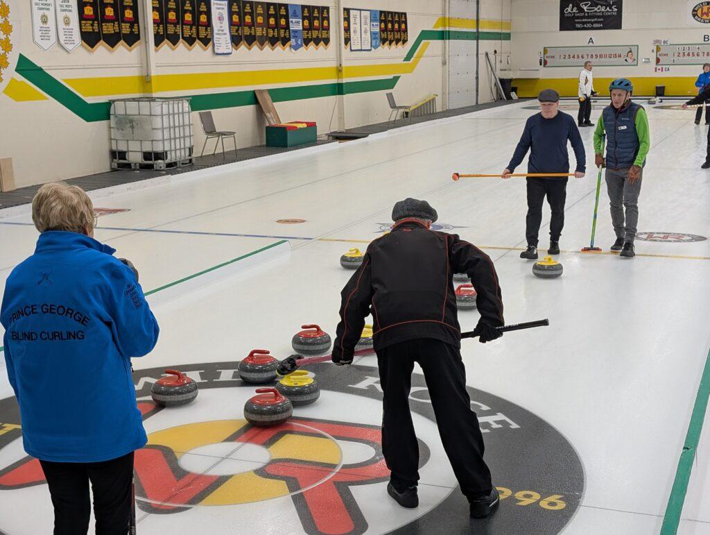 Players from Team Canada can be seen playing against Team British Columbia holding brooms. Red and yellow curling stones are on the ice.