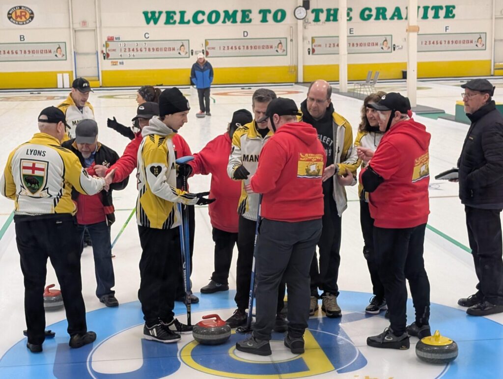 Team Manitoba players wearing their yellow and white team jackets shake hands with players from Team New Brunswick players wearing their red team jackets on the ice following the end of the consolation final. Yellow and red curling stones are scattered around them.