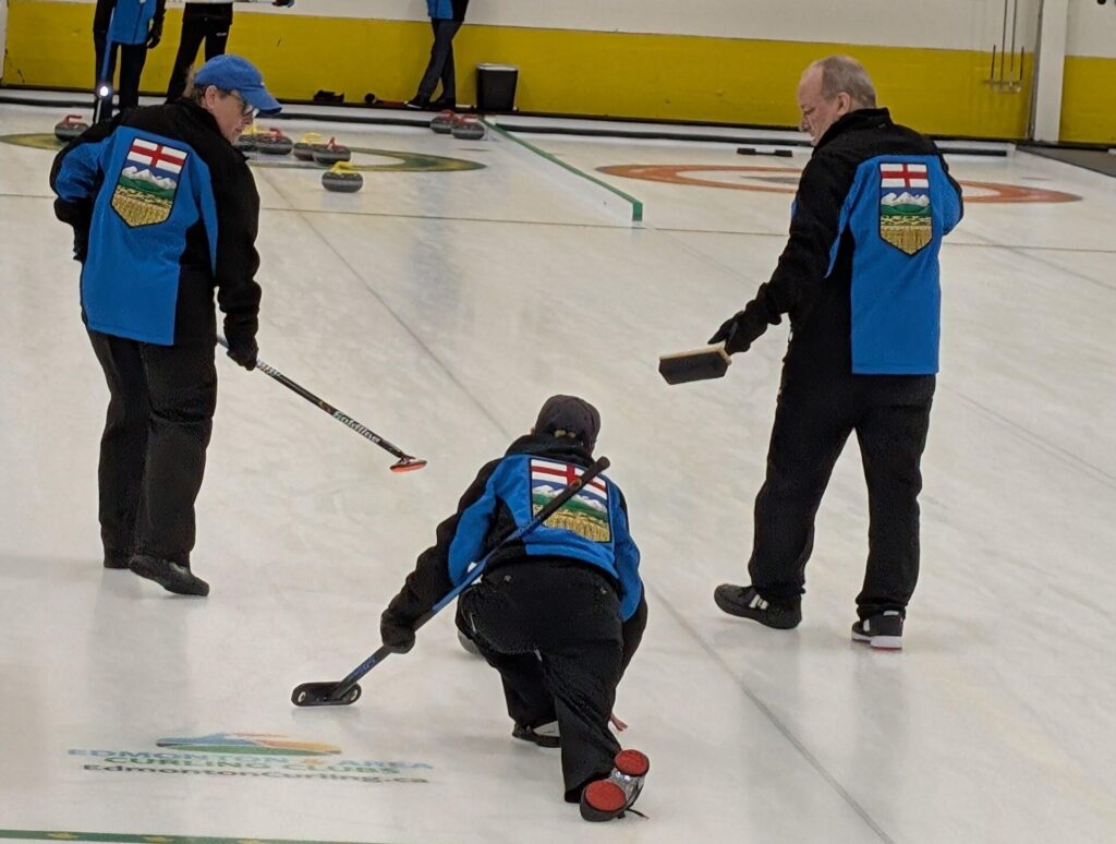 Players from Team Alberta, wearing blue and black jackets with the Alberta flag on the back, are competing against Team British Columbia in the gold medal game. One player is delivering a curling stone while teammates sweep the ice.