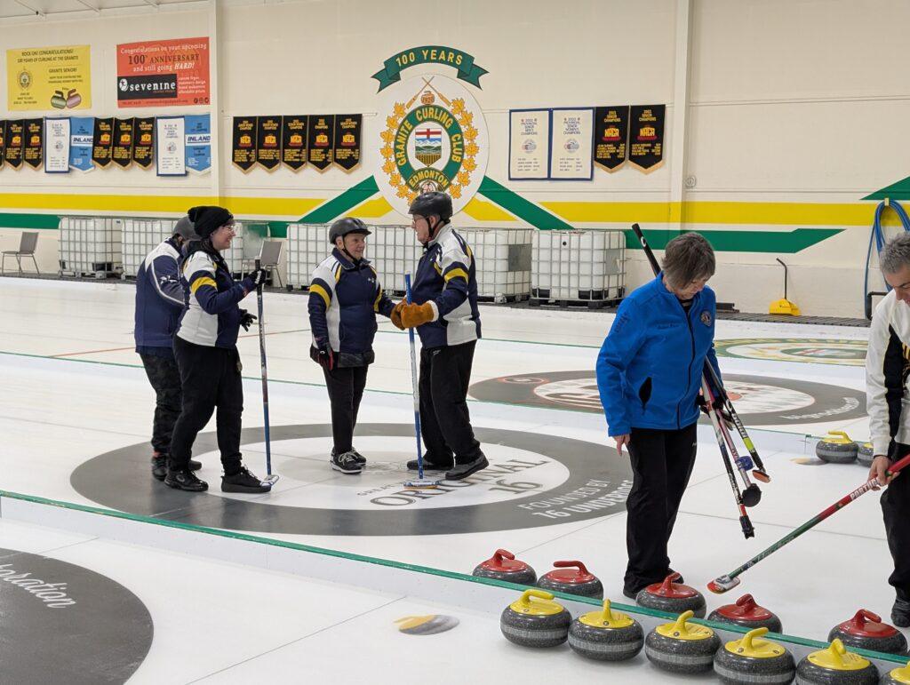Team Nova Scotia players in dark blue and yellow jackets are talking to each other on the ice, with Team Canada players in blue and white jackets nearby next to lined-up red and yellow curling stones. Players are holding curling brooms.