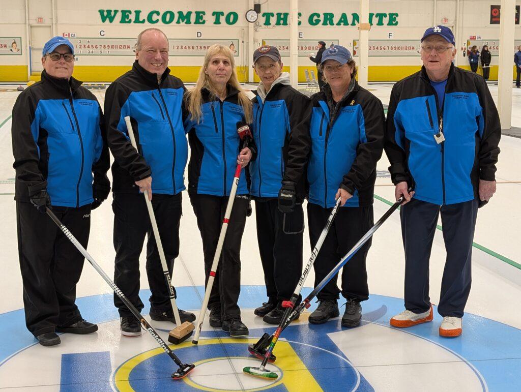 Team Alberta players stand on the ice wearing their blue and black team jackets, each holding a broom and smiling for the camera.