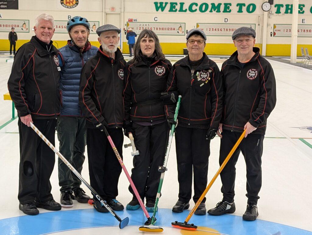 Team British Columbia players stand on the ice wearing their black and red team jackets, each holding a broom and smiling for the camera. One player wears a dark blue vest.