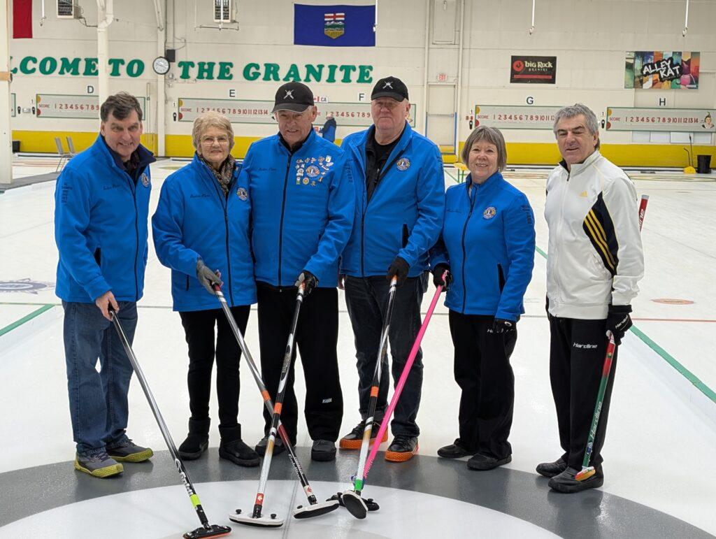 Team Canada (BC) players stand on the ice wearing their blue team jackets with one player in a white jacket, each holding a broom and smiling for the camera.