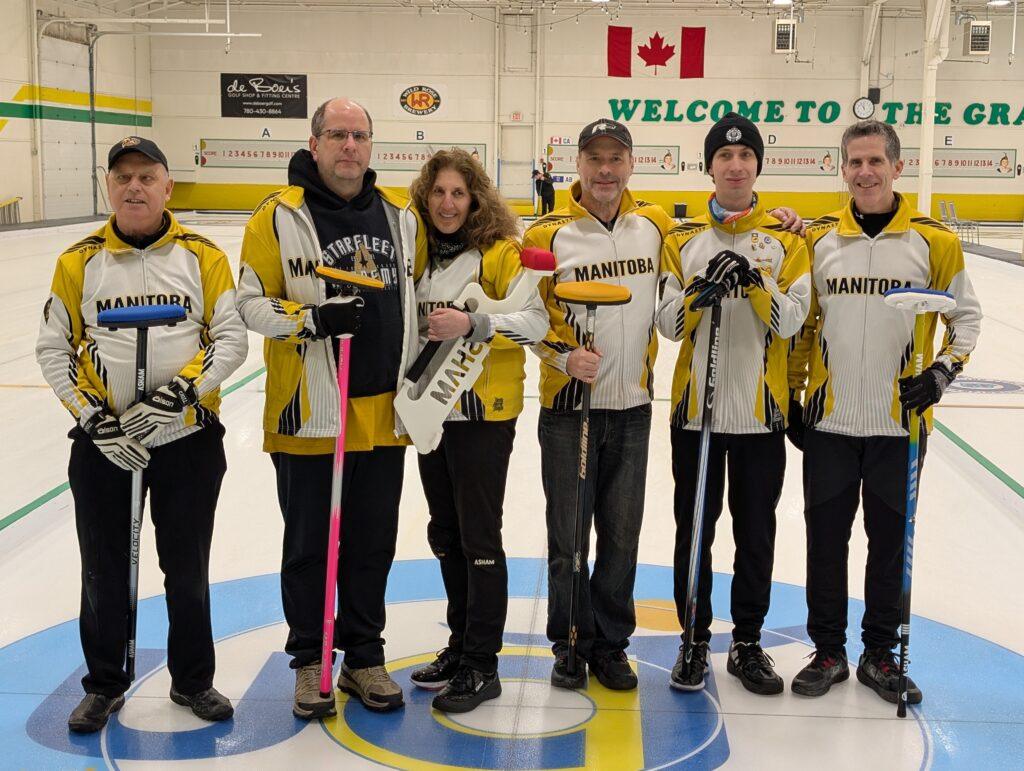 Team Manitoba players stand on the ice wearing their yellow and white team jackets, most holding a broom and smiling for the camera.