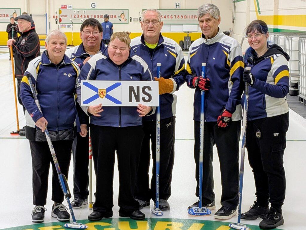 Team Nova Scotia players stand on the ice wearing their dark blue, yellow, and white team jackets, each holding a broom and smiling for the camera. One player holds a NS sign with the Nova Scotia flag.