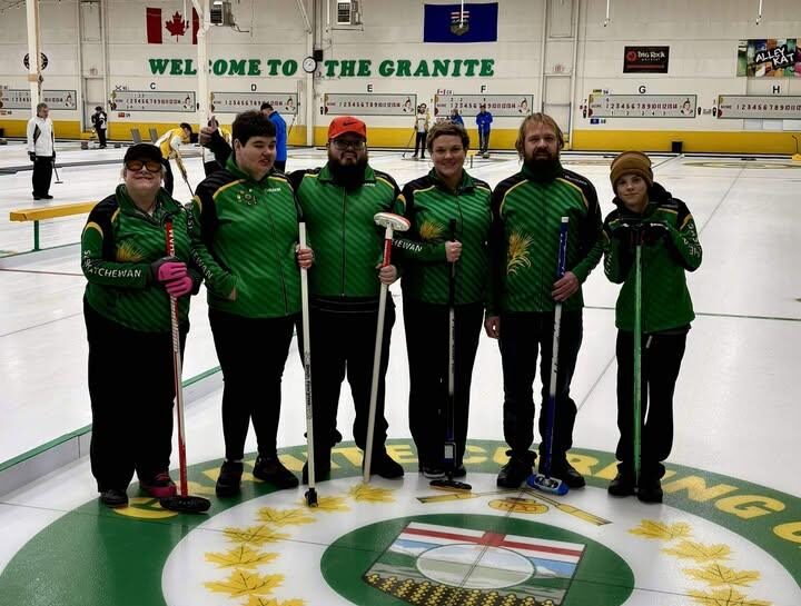 Team Saskatchewan players stand on the ice wearing their green, black, and yellow team jackets, each holding a broom and smiling for the camera.