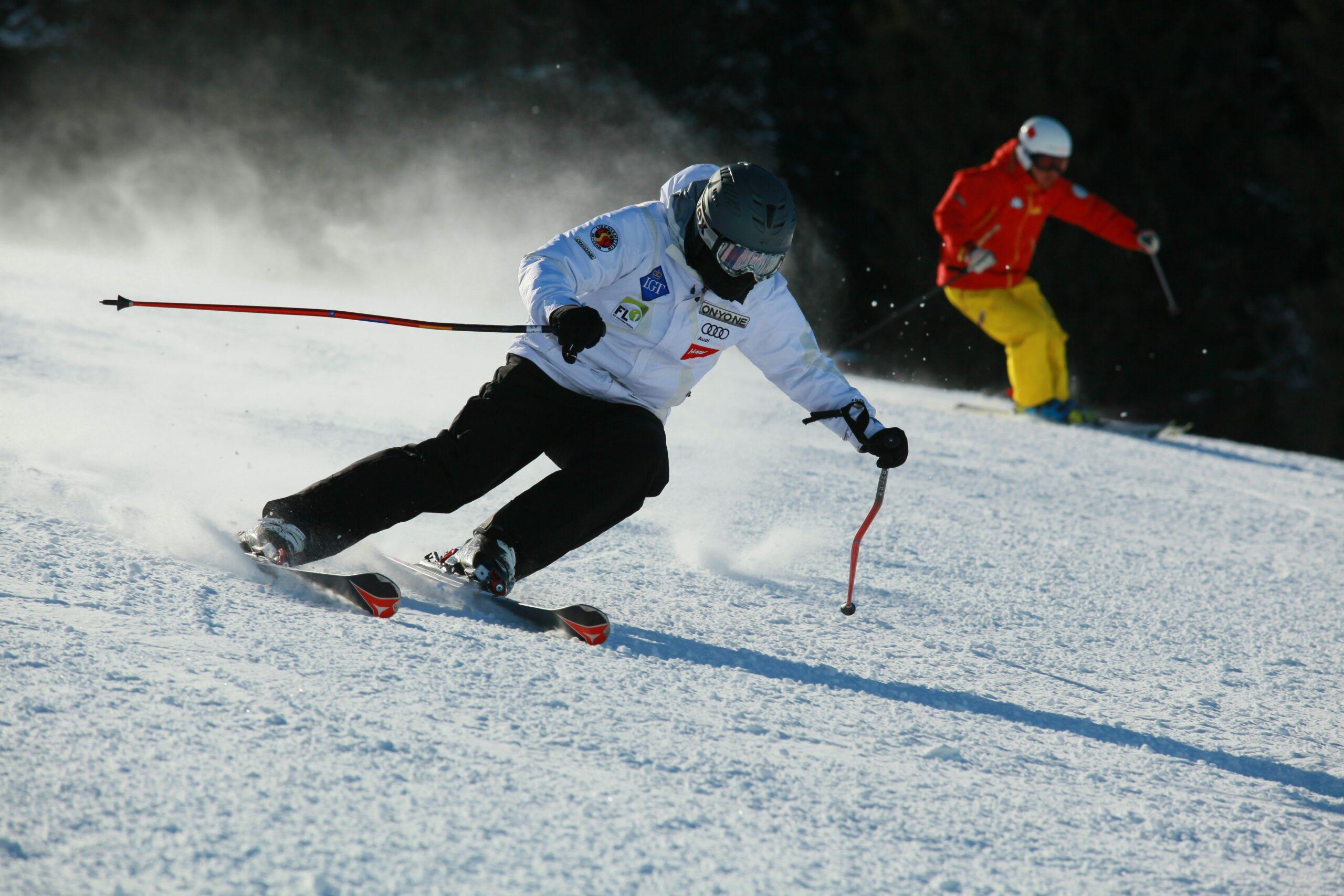 A skier in a white jacket and black snow pants carves sharply down a snowy slope while another skier in a red jacket and yellow snow pants follows in the background.