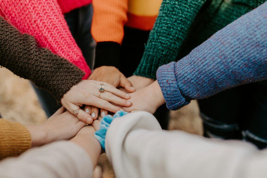 A group of women place their hands together in a stack, each wearing long-sleeve sweaters in different colours.