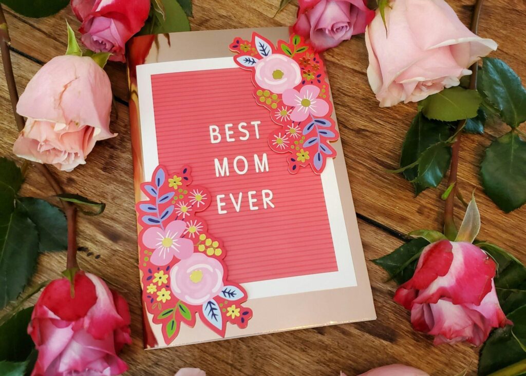 A card with coral pink, blue, green, and yellow floral designs lays flat on a wooden table surrounded by light and dark pink roses. White text on the cover of the card reads: Best Mom Ever.