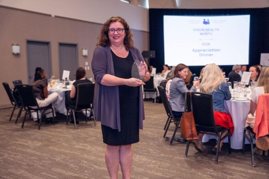 Jennifer Urosevic, President of Vision Loss Rehabilitation Canada, stands smiling while holding the Canadian Council of the Blind’s Person of the Year award. She is wearing a black dress with a dark purple cardigan and poses proudly with the award in hand. People are seated at tables behind her, with a presentation slide displayed on a screen in the background.