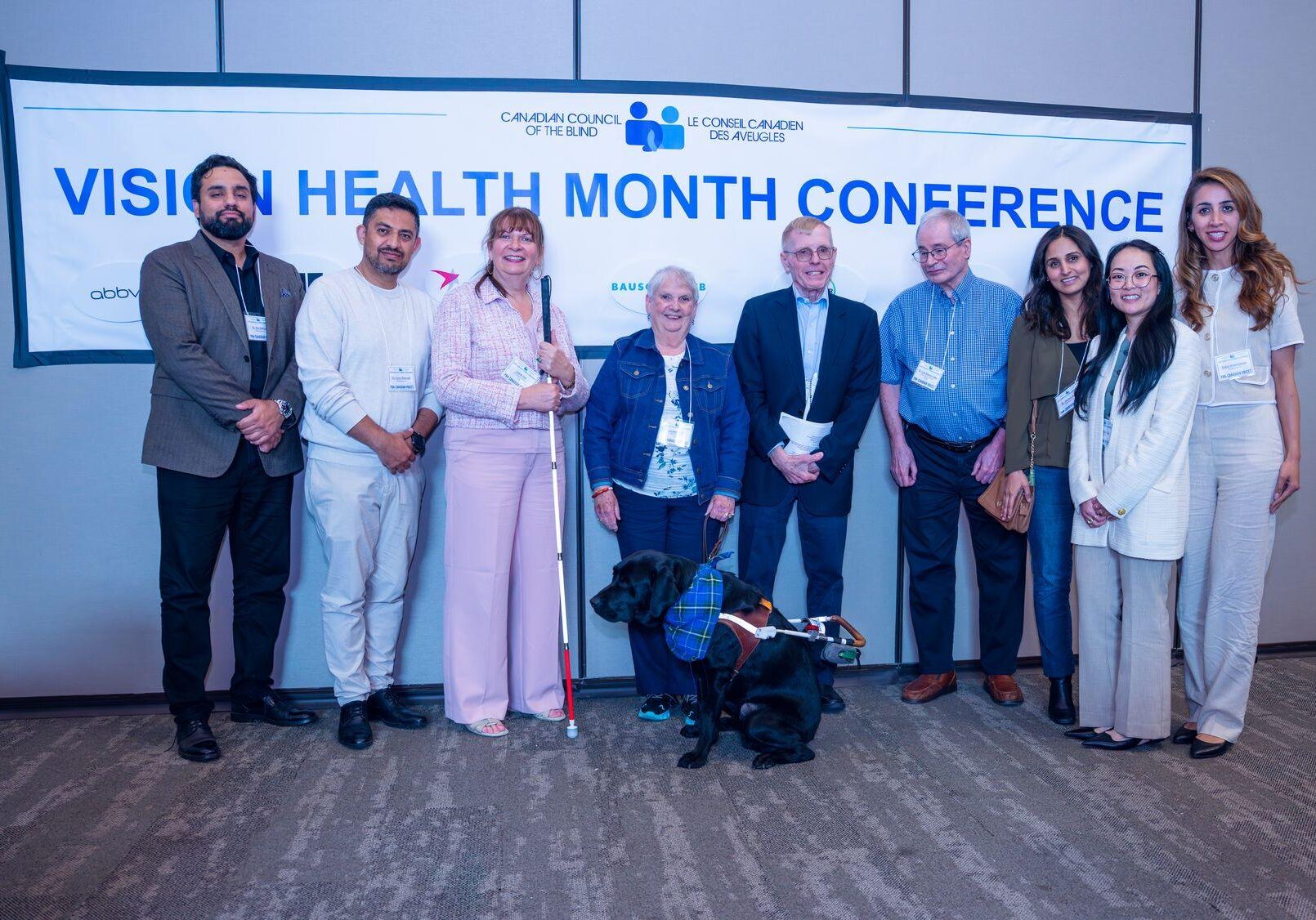 Individuals representing Specsavers smile and pose for a group photo, with CCB’s Leslie Yee, Louise Gillis and her guide dog, Monk, Jim Tokos, and Leo Bissonnette in the center. Behind them is a banner displaying the CCB logo and the words “Vision Health Month Conference” in blue on a white background.