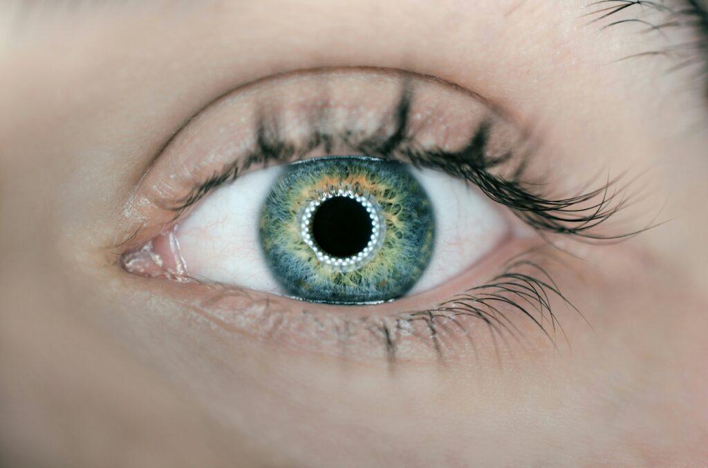 Close-up of an eye in shades of turquoise, blue, green, light yellow, and orange. A ring of lights can be seen around the black pupil.