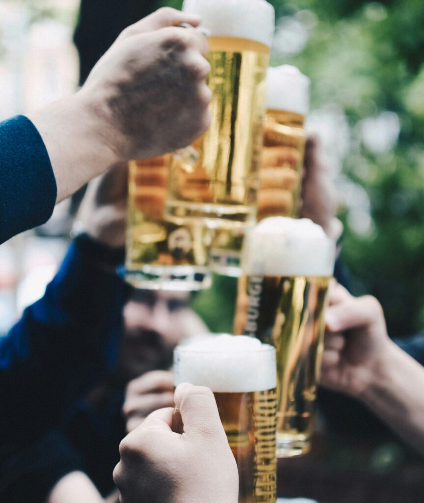 Close-up of hands raising glasses of beer with greenery visible in the blurred background.