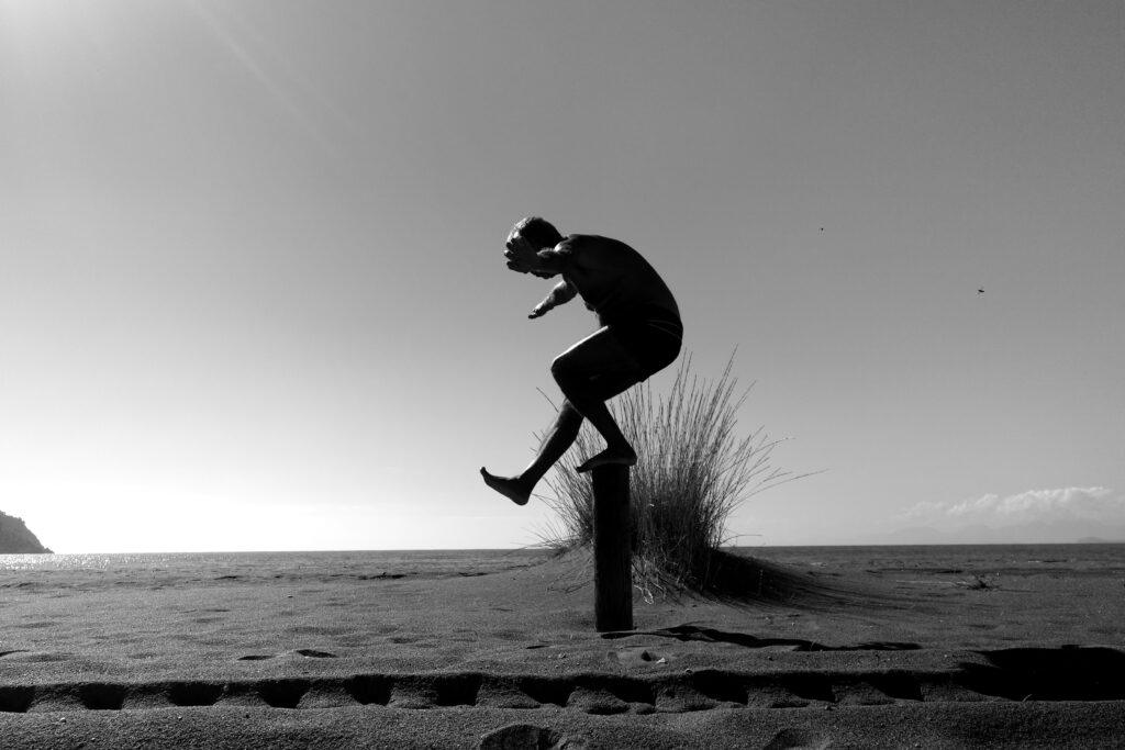 A grayscale image of an individual balancing on one leg with their arms raised on a small pillar in the sand. The ocean can be seen behind.