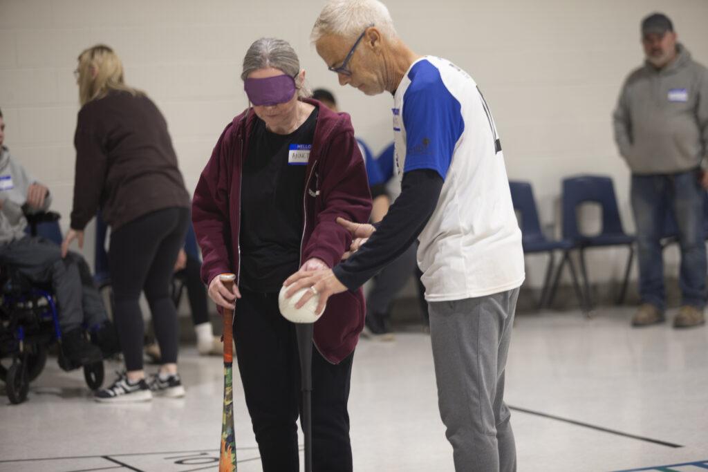 A photo of a woman and a man in the center of a room, with other people and chairs along the wall in the background. The woman is blindfolded and holds a white ball in her left hand and a baseball bat in her right hand. The man beside her, wearing a white shirt with short blue sleeves over a long-sleeve black shirt, is guiding her by holding her left arm and helping position the ball.