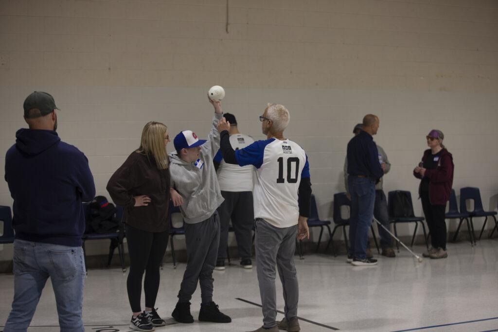 A photo of a group of people, with two individuals centered in the frame. One is wearing a baseball cap, raising his left arm while holding a white ball. The other is wearing a white shirt with short blue sleeves over a long-sleeve black shirt, with the number 10 on the back.