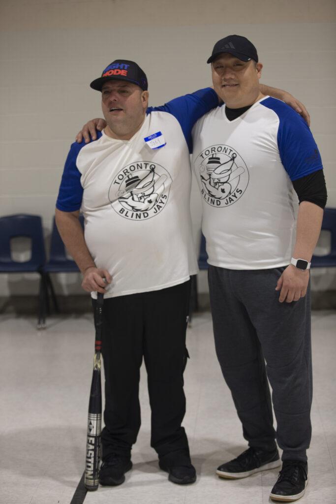Two men stand posing for the camera, each with an arm around the other’s shoulder. The man on the right is holding a baseball bat in his right hand. Both are wearing white shirts with short blue sleeves and a circular logo in the center that reads ‘Toronto Blind Jays.’ Both are also wearing black baseball caps.