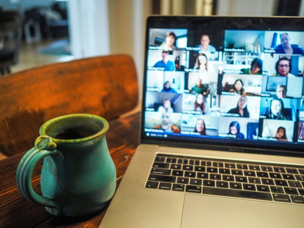 On a wooden table, a large turquoise mug sits on the left, and an open laptop is on the right during a Zoom call. Several participants appear in small rectangles on the laptop screen.