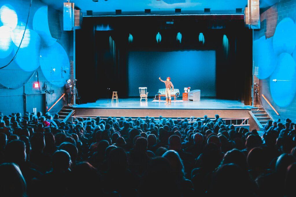 The backs of audience members are visible in the foreground, facing a stage where a person is performing. The stage is illuminated with blue lighting.