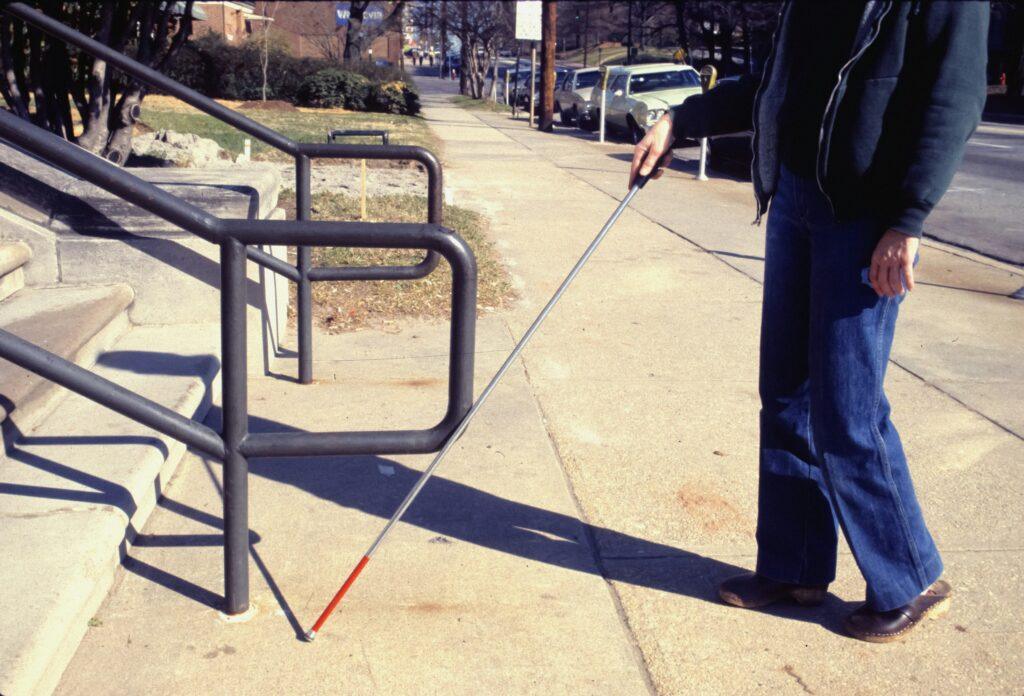 A person is shown from the mid-arm down, wearing a grey jacket and blue jeans, using a white cane while approaching a flight of stairs with grey metal railings. They are outdoors on a paved surface on a bright, sunny day, with cars parked along the side of the road.