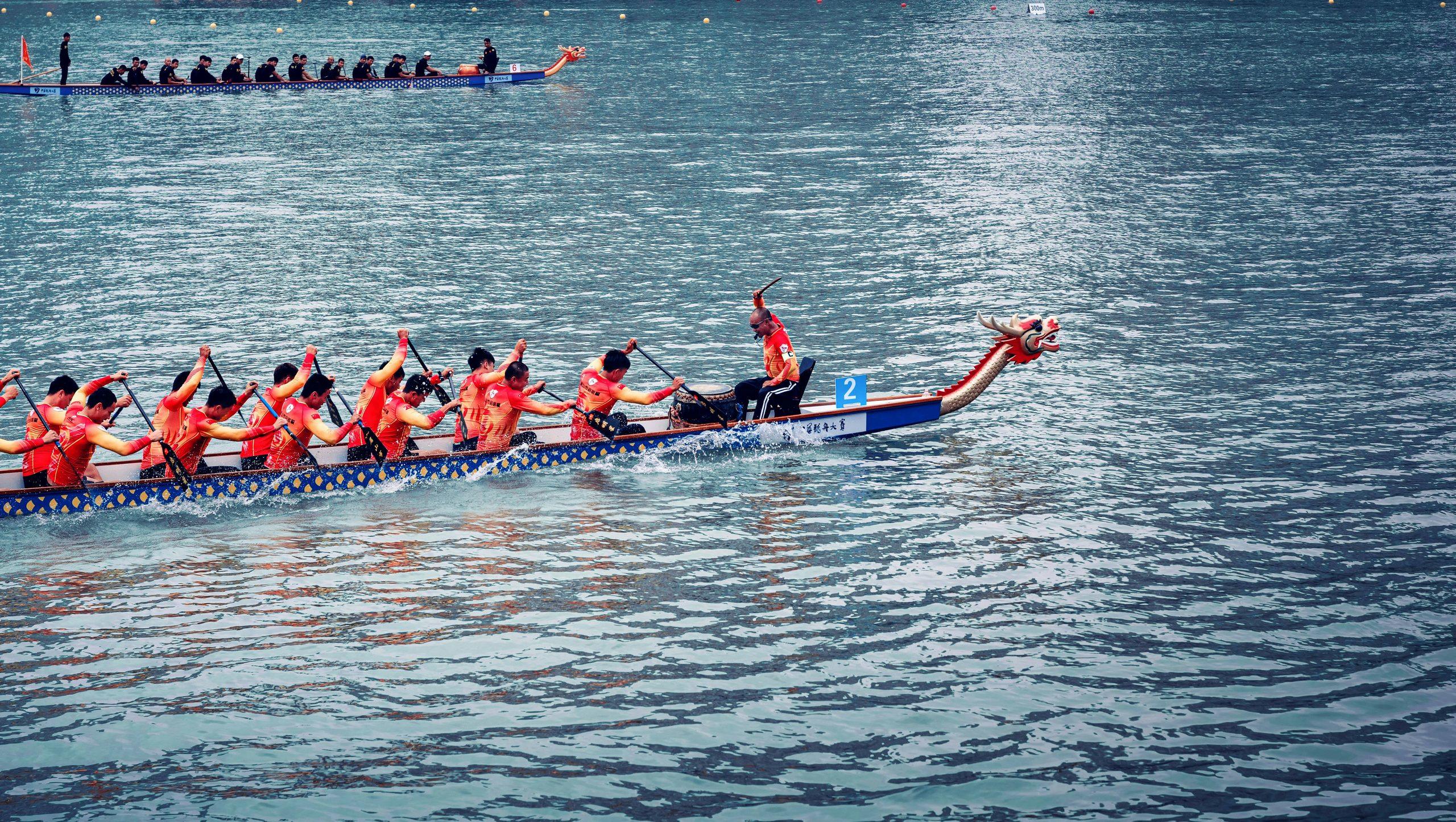 Several people wearing bright orange shirts paddle two by two in a blue dragon boat decorated with orange spots. A dragon head is attached to the front of the boat. At the front, a person sits holding a baton, poised to strike a drum. Another dragon boat can be seen in the distance alongside them.