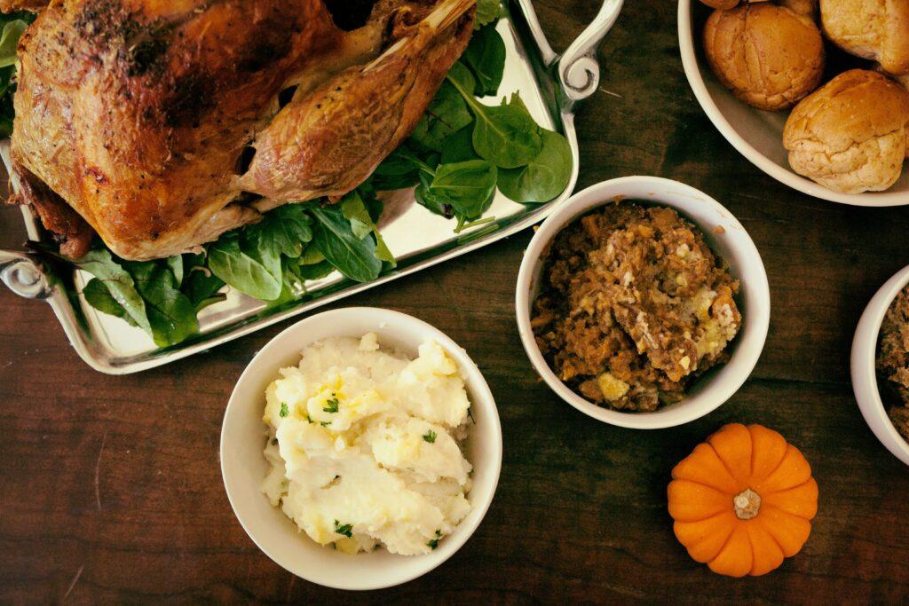 Overhead view of a cooked turkey above spinach leaves on a silver pan, mashed potatoes, stuffing, and dinner buns in white bowls, with a small decorative orange pumpkin, all arranged on a wooden table.